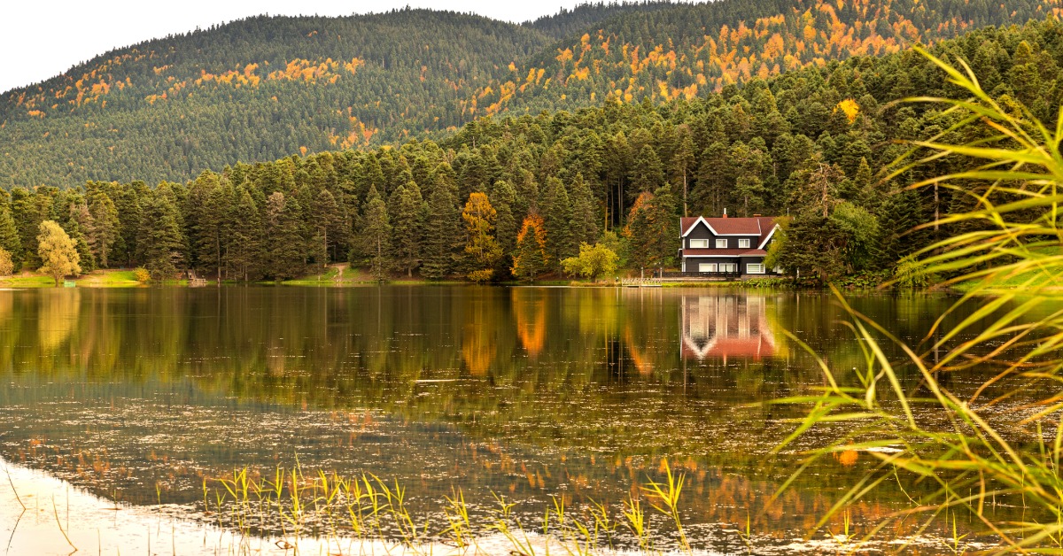 A lakeside house sits across calm water, surrounded by forested hills with autumn foliage reflected on the surface.