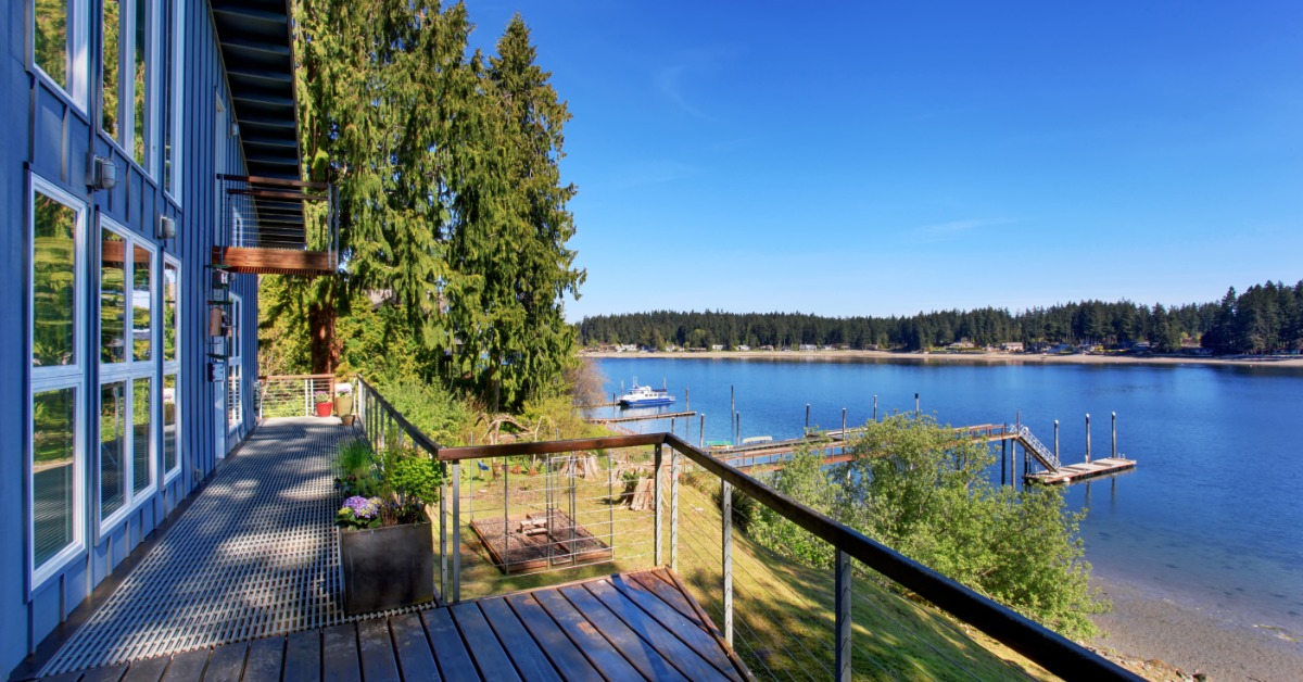 A lakeside home features a deck overlooking calm water, with docks, trees, and a clear blue sky in view.