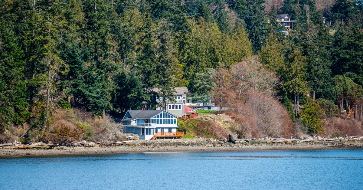 A waterfront house sits along a rocky shoreline, surrounded by trees, with calm water in the foreground.