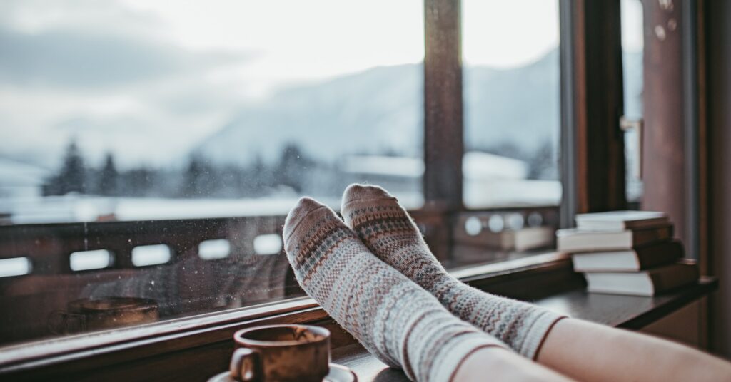 A person wearing cozy knit socks relaxes by a window with a warm drink while snowy mountains stretch into the distance.