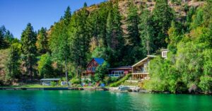 A lakeside home sits beneath forested mountains with private docks overlooking clear green water on a sunny day.