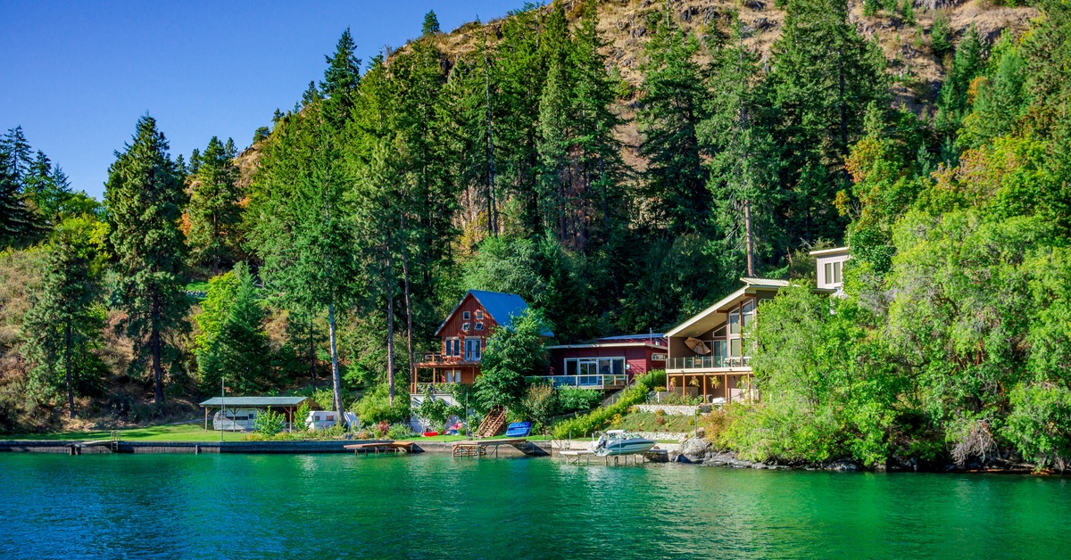 A lakeside home sits beneath forested mountains with private docks overlooking clear green water on a sunny day.