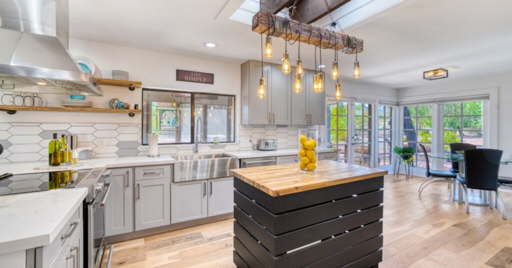 A bright modern kitchen features a large island, skylight, pendant lighting, and windows overlooking a wooded yard.