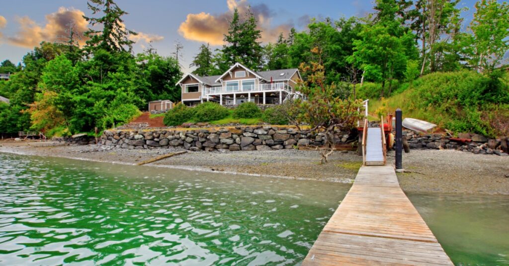 A wooden dock leads to a waterfront home surrounded by trees along a calm shoreline beneath an evening sky.