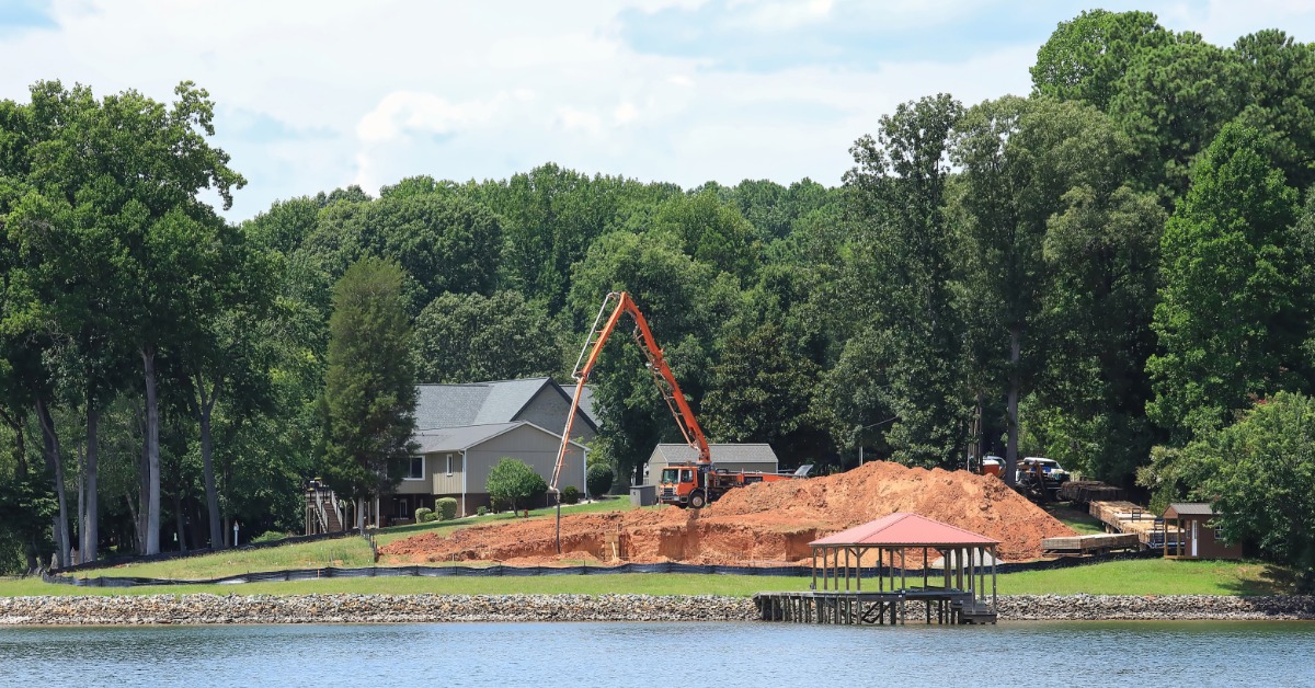 A concrete pump truck pours a home's foundation beside a lakeshore surrounded by mature trees on a sunny day.