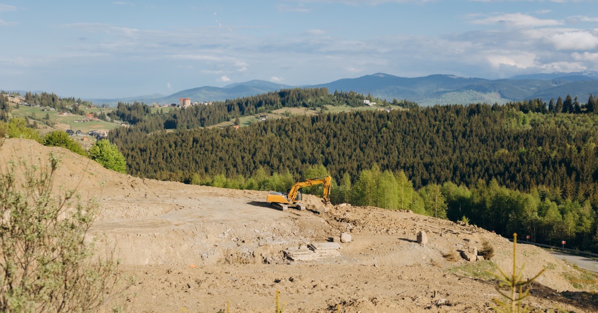 An excavator clears a hillside terrain construction site overlooking forested mountains beneath blue skies.