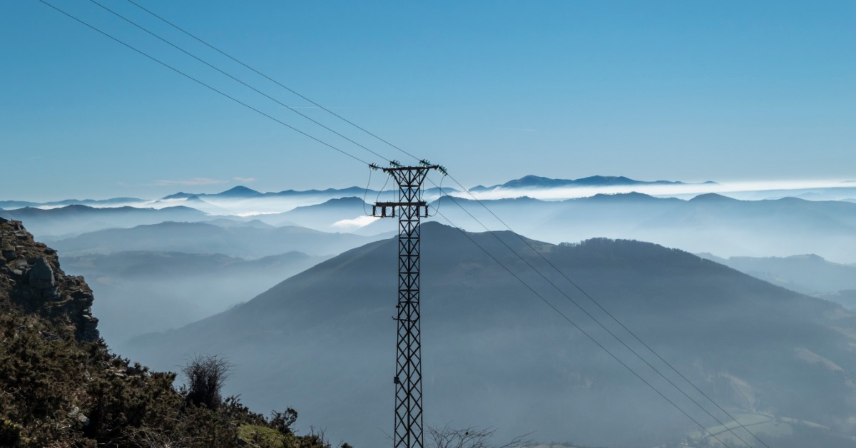 A power line tower stands on a mountain ridge, with cables stretching across layered hills under a clear blue sky.