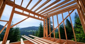 A wooden house frame under construction overlooks a forested mountain landscape with clear skies in the background.