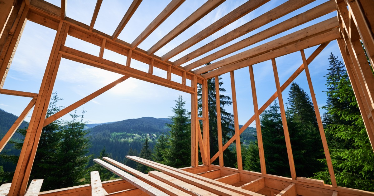 A wooden house frame under construction overlooks a forested mountain landscape with clear skies in the background.