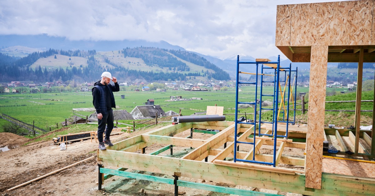 A construction worker stands on a raised foundation at a mountain building site overlooking a rural valley and hills.