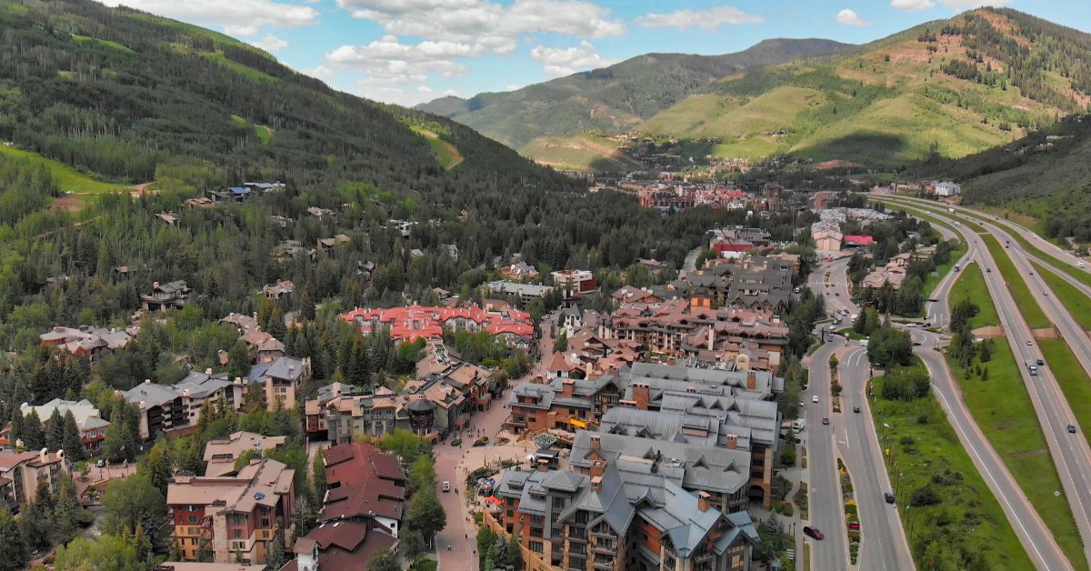 An aerial view shows a mountain town with homes, roads, and resort buildings surrounded by forested slopes.