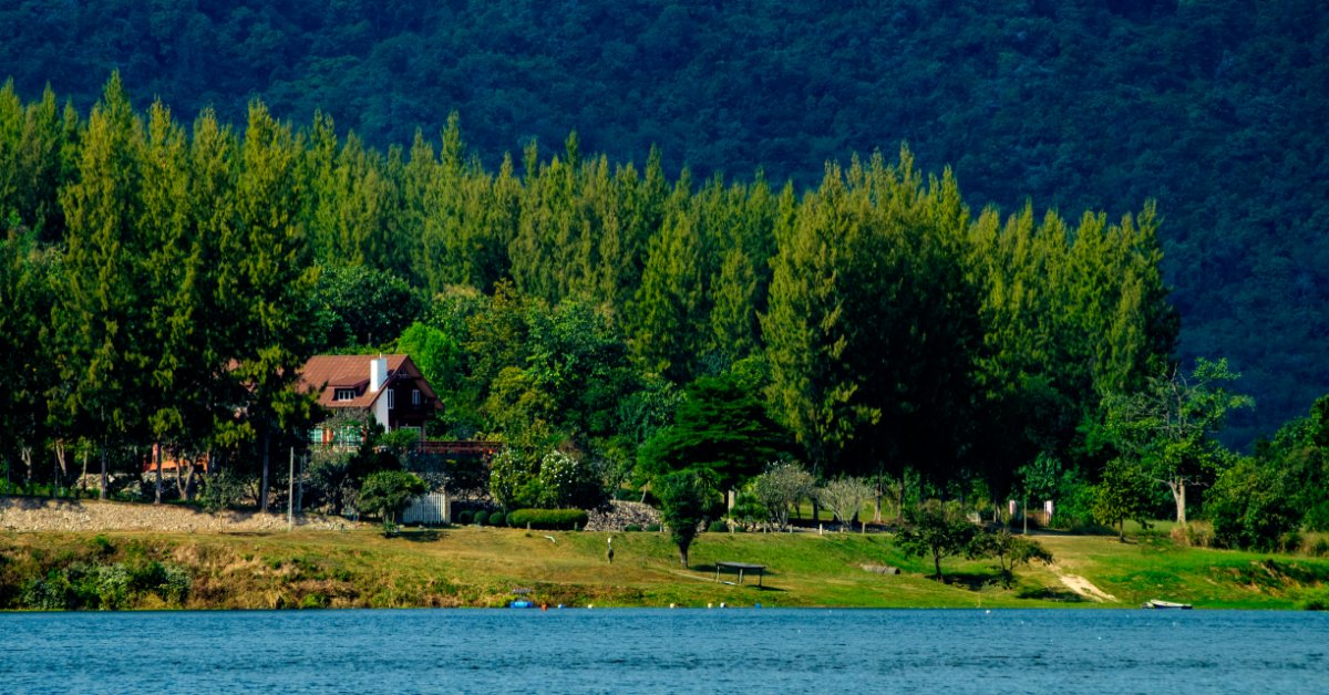 A large two-story home sits among dense trees beside a lake, with forested mountains in the background.