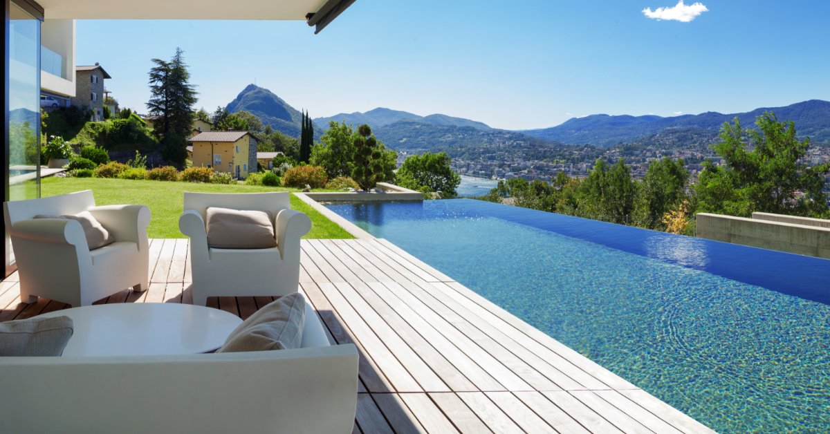A mountain home patio with an infinity pool overlooks a lake, a nearby town, and the surrounding mountain landscape.