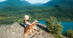 A hiker sits with a golden retriever on a rocky overlook above a blue lake and forested mountains below.