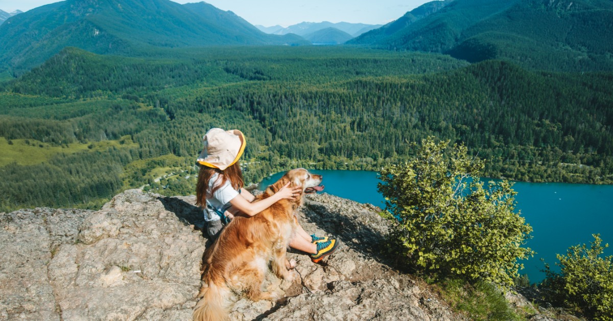 A hiker sits with a golden retriever on a rocky overlook above a blue lake and forested mountains below.