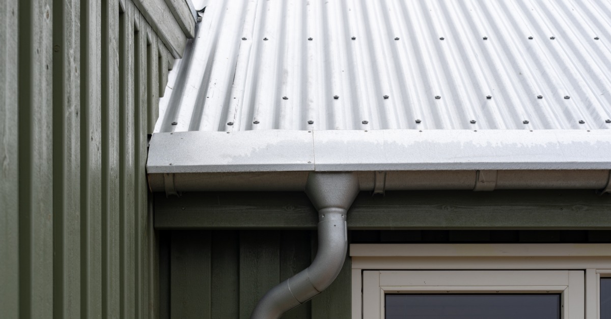 A metal roof with a gutter and downspout channels water along the green exterior of a residential home.