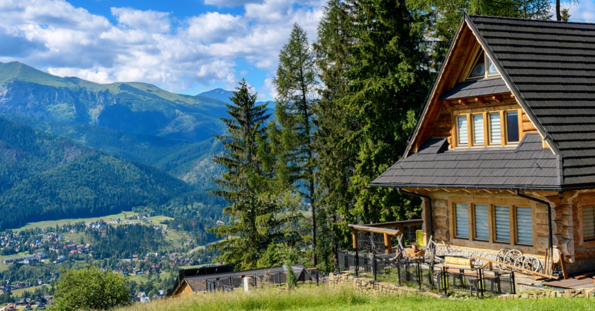 A wooden mountain home overlooks a forested valley and distant peaks beneath a bright, partly cloudy sky.