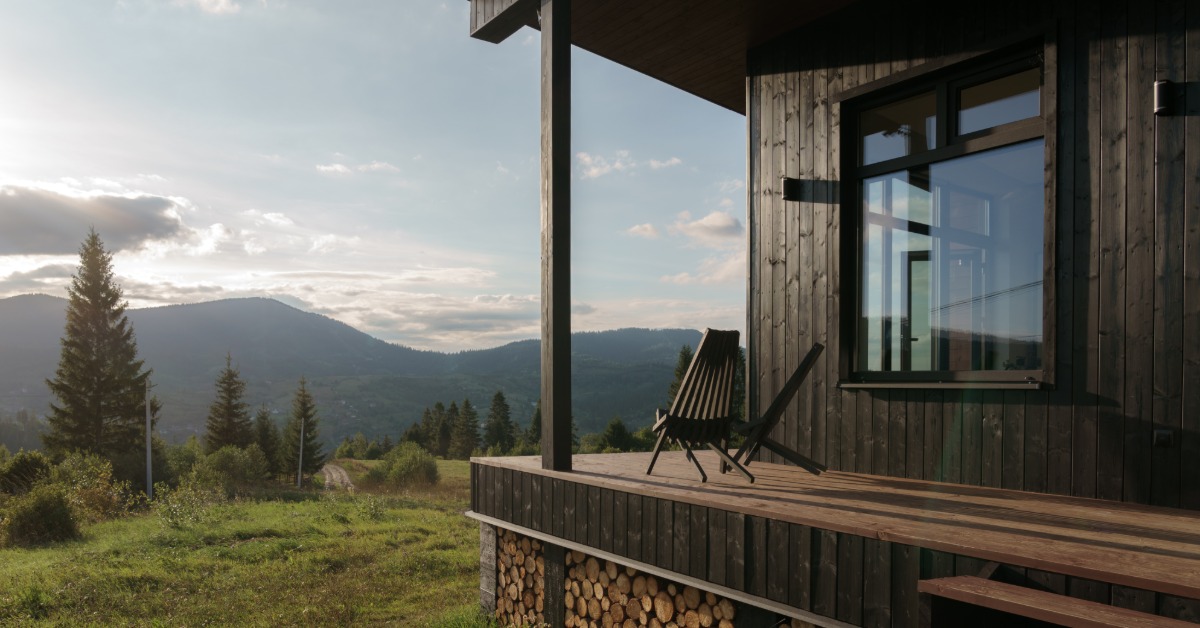 A mountain home with a wooden deck and stacked firewood overlooks a forested valley and distant peaks.