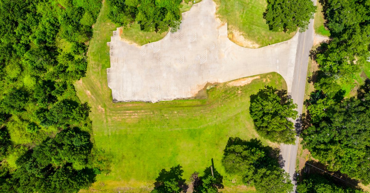 An aerial view shows a vacant grassy property bordered by two paved roads and surrounded by dense green trees.